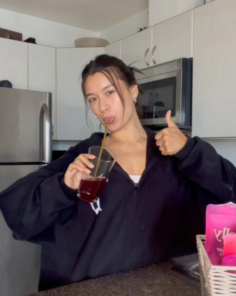 A person in a black hoodie stands in a kitchen, holding a glass of red liquid with a straw. They are giving a thumbs-up with their other hand. A basket and a pink item are visible on the counter.